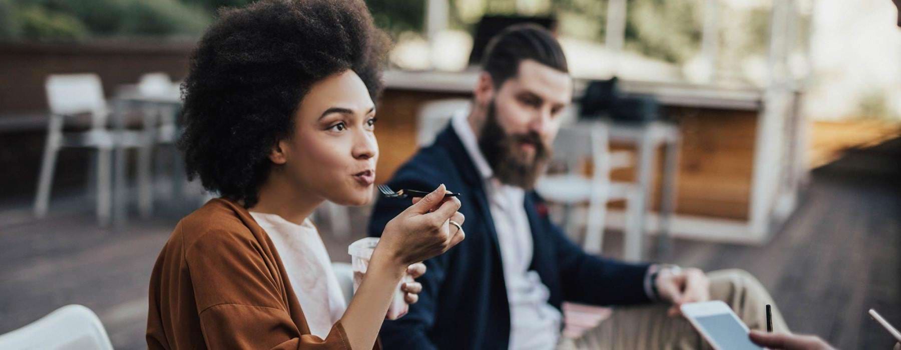 woman sits and eats outside while someone holding a cell talks to her